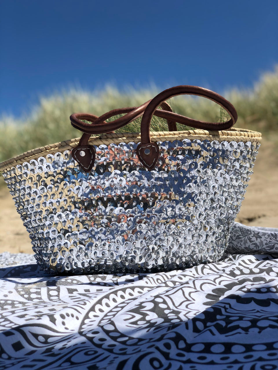 Large straw beach bag with sequins silver, on a beach blanket under a blue sky
