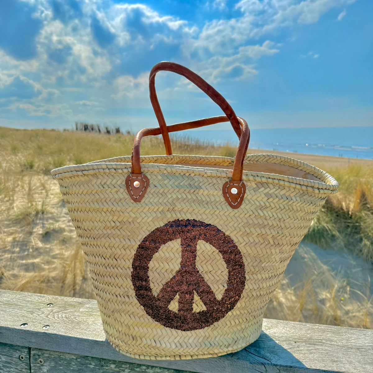 Rieten strandtas met pailletten Peace bruin, in de duinen onder een blauwe lucht